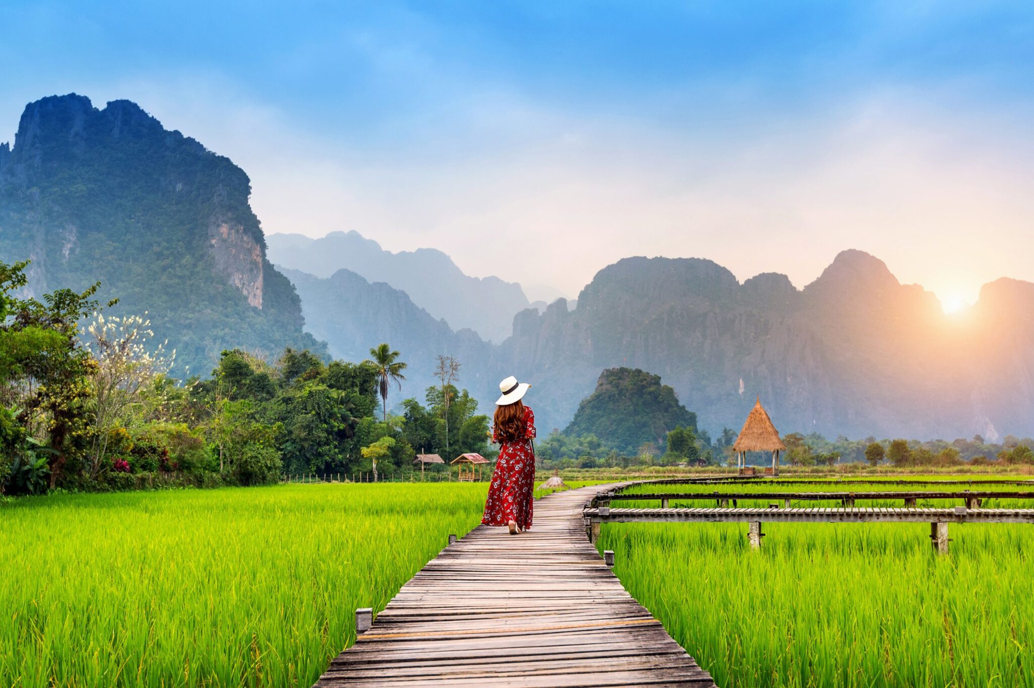 young-woman-walking-wooden-path-with-green-rice-field-vang-vieng-laos-scaled